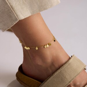 May include: A gold-colored anklet with small, round charms is worn on an ankle. The ankle is adorned with a delicate chain and several disc-shaped pendants. The person is wearing beige sandals with a light-colored sole.
