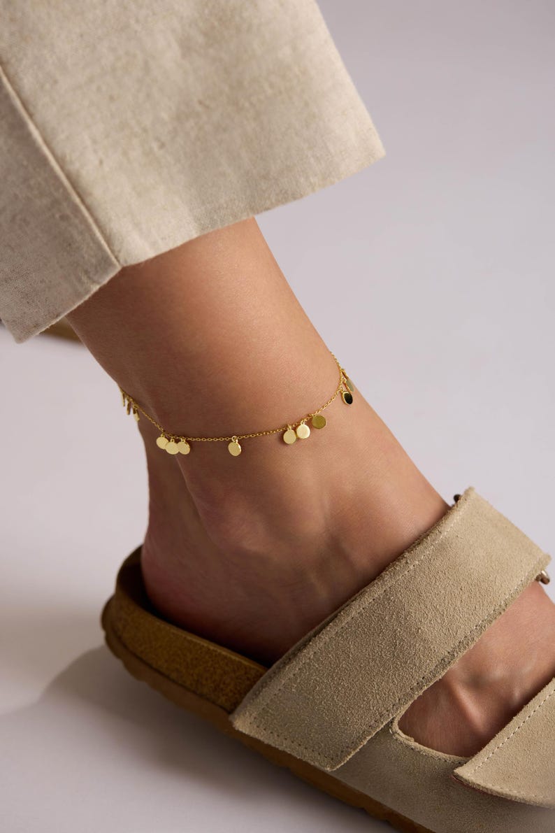 May include: A gold-colored anklet with small, round charms is worn on an ankle. The ankle is adorned with a delicate chain and several disc-shaped pendants. The person is wearing beige sandals with a light-colored sole.