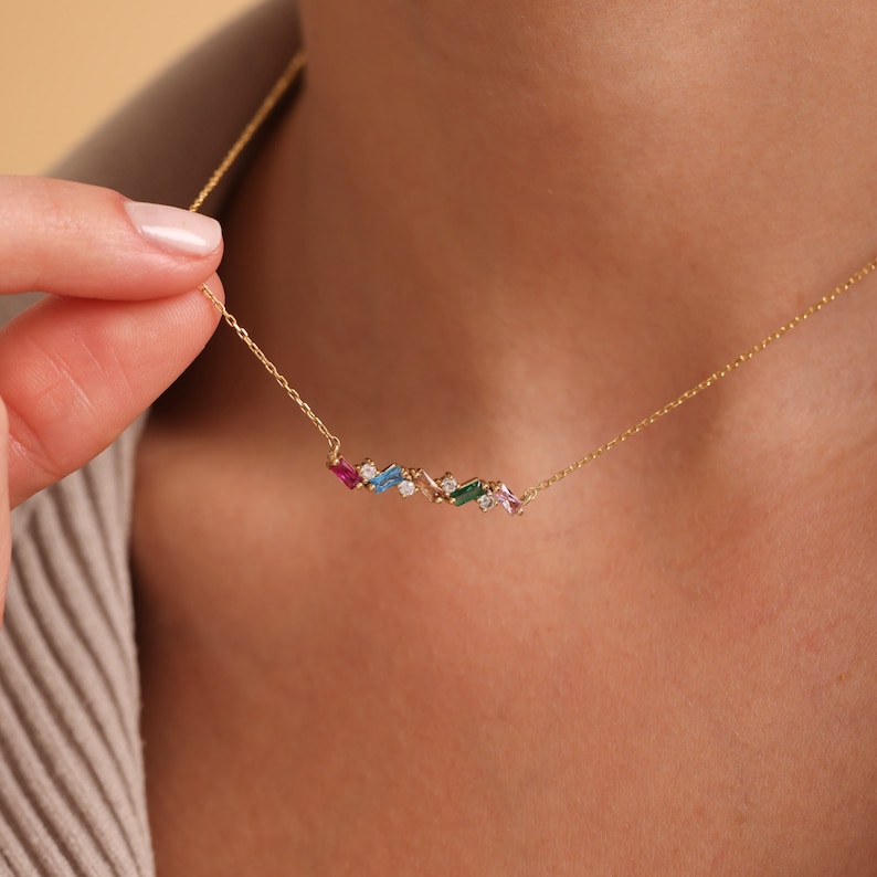 Puede incluir: Un collar de cadena de oro con un colgante de barra de piedras preciosas de colores. La barra presenta piedras preciosas rosas, azules, verdes y transparentes.