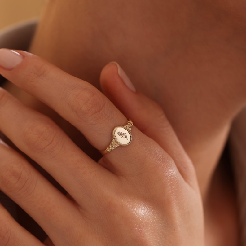 May include: A gold signet ring with an oval shaped top featuring a delicate leaf design.