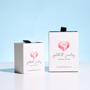May include: Two white jewelry boxes with black ribbon pulls. Each box features a geometric pink heart and the words "goldhill jewelry handmade jewelry" in black script. The website address is printed below.