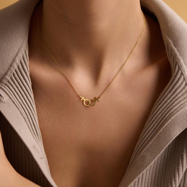 May include: A gold necklace with a delicate chain and a pendant featuring interlocking female and male symbols.