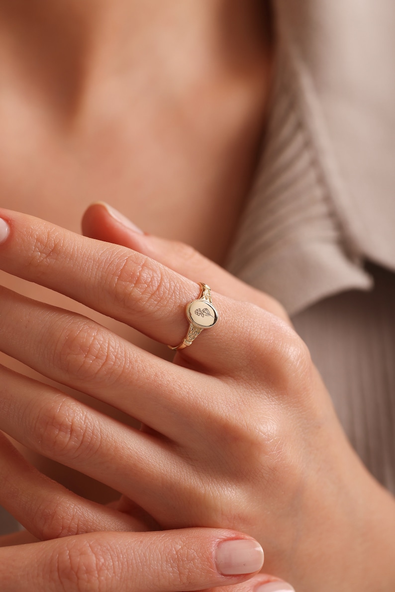 May include: A gold signet ring with a floral design on the face. The ring is worn on the index finger of a hand.