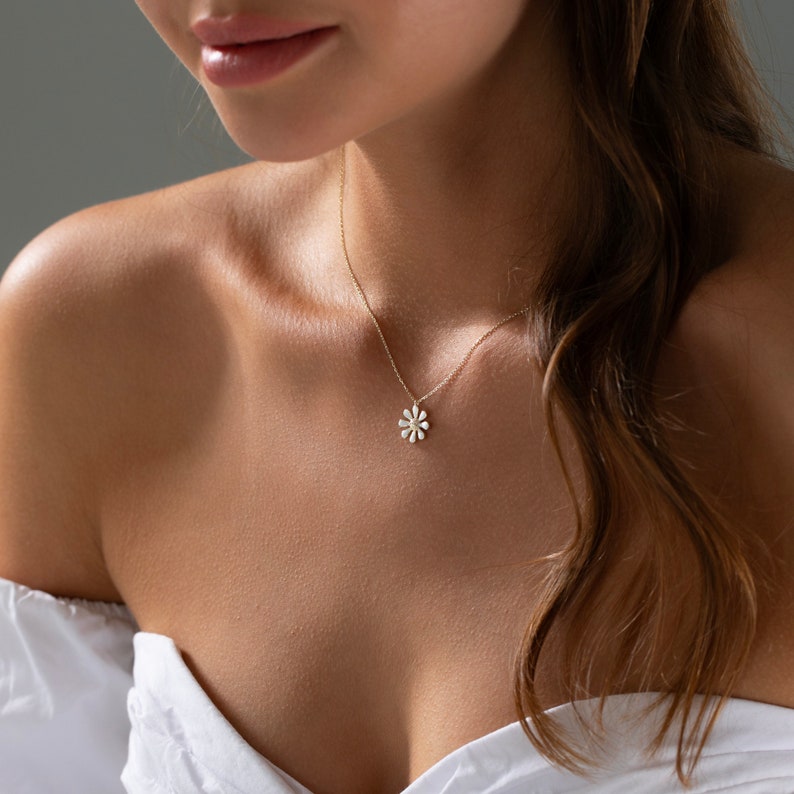 May include: A delicate gold necklace with a small, white daisy pendant. The necklace is worn by a person with long, brown hair.