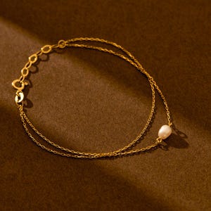 May include: Delicate gold-toned bracelet featuring a double chain design. A single, lustrous white pearl is suspended from the lower chain. The bracelet is set against a warm brown backdrop, highlighting its elegant simplicity.