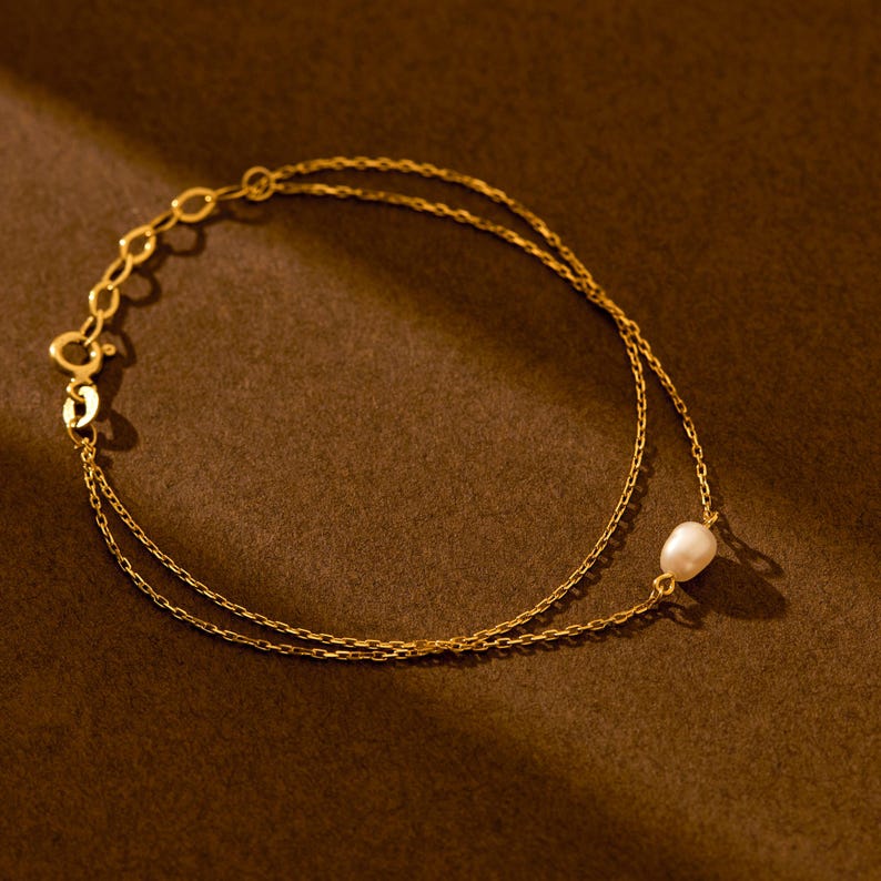May include: Delicate gold-toned bracelet featuring a double chain design. A single, lustrous white pearl is suspended from the lower chain. The bracelet is set against a warm brown backdrop, highlighting its elegant simplicity.