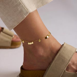 May include: A gold-colored anklet with small, round disc charms. The delicate chain anklet is worn on an ankle with a sandal. The anklet's charms are evenly spaced along the chain.