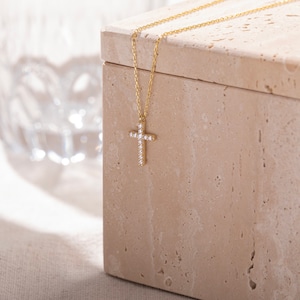 K&ouml;nnte beinhalten: Eine goldene Kettenkette mit einem kleinen, diamantbesetzten Kreuzanh&auml;nger. Die Kette wird auf einer beigefarbenen Steinkiste pr&auml;sentiert.