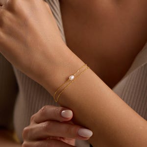 May include: A delicate gold bracelet with a small, round pearl at the center. The bracelet is a double-strand design, and the gold chain is thin and elegant. The bracelet is worn on a wrist, and the skin tone is light.