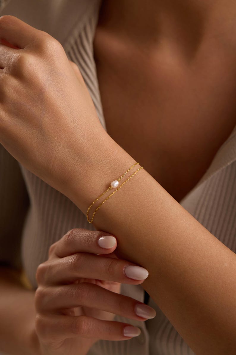 May include: A delicate gold bracelet with a small, round pearl at the center. The bracelet is a double-strand design, and the gold chain is thin and elegant. The bracelet is worn on a wrist, and the skin tone is light.