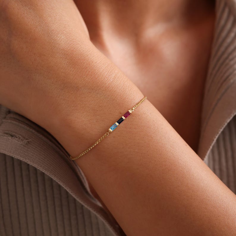 May include: A gold chain bracelet with four small, colorful gemstones. The gemstones are blue, dark blue, red, and yellow.