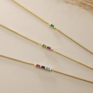 May include: Three gold chain necklaces with different colored gemstones. The top necklace has a green gemstone, the middle necklace has a green, pink, and yellow gemstone, and the bottom necklace has a pink, blue, and light blue gemstone.