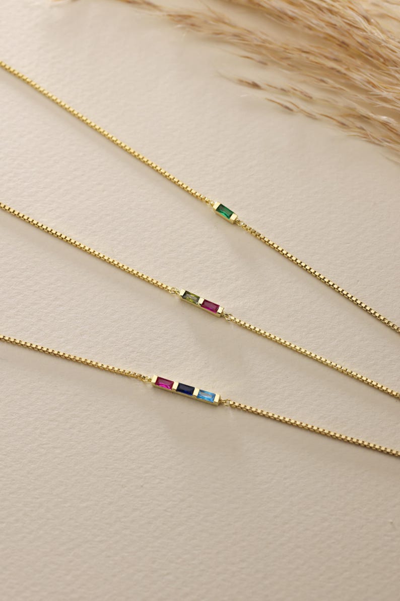 May include: Three gold chain necklaces with different colored gemstones. The top necklace has a green gemstone, the middle necklace has a green, pink, and yellow gemstone, and the bottom necklace has a pink, blue, and light blue gemstone.