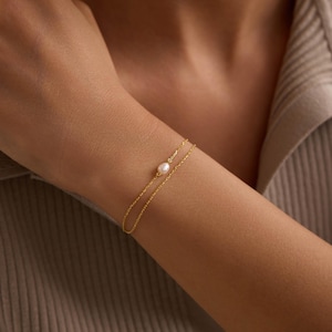 May include: A delicate gold bracelet featuring a small, round pearl. The bracelet has a double-strand design and is worn on a person's wrist. The jewelry is simple and elegant, suitable for everyday wear.