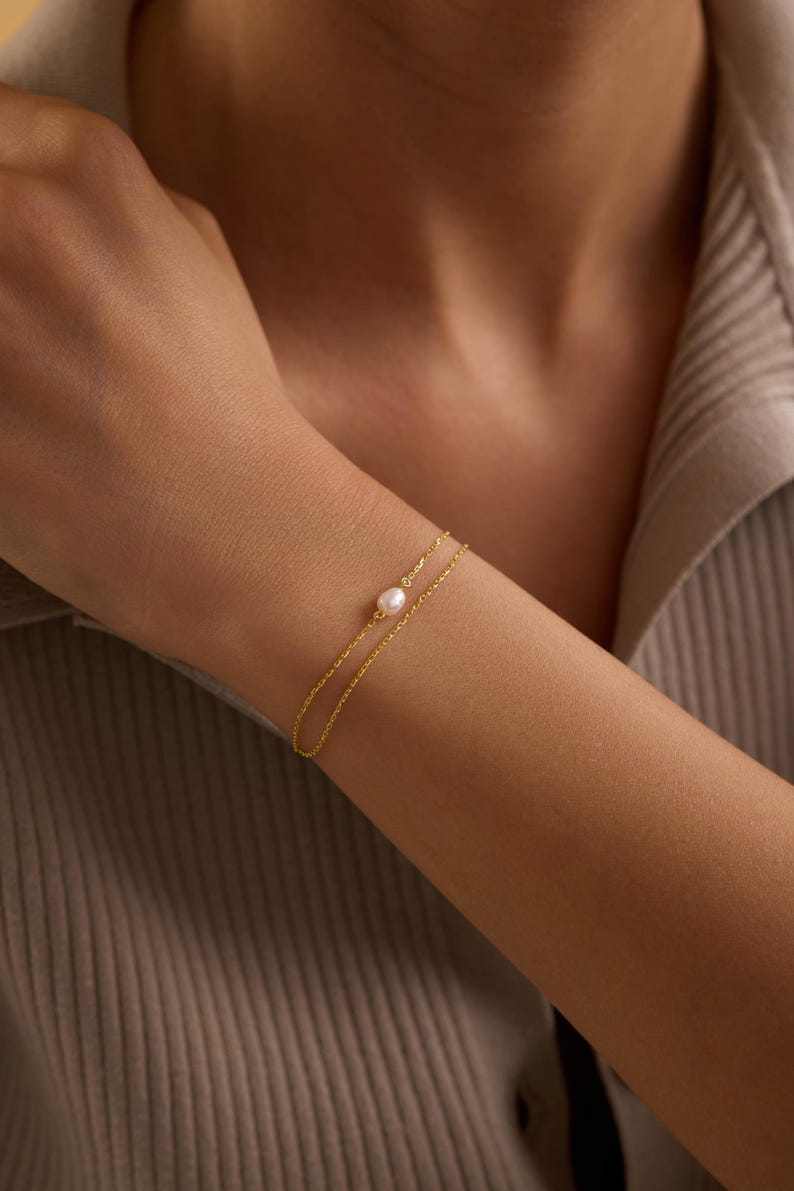 May include: A delicate gold bracelet featuring a small, round pearl. The bracelet has a double-strand design and is worn on a person's wrist. The jewelry is simple and elegant, suitable for everyday wear.