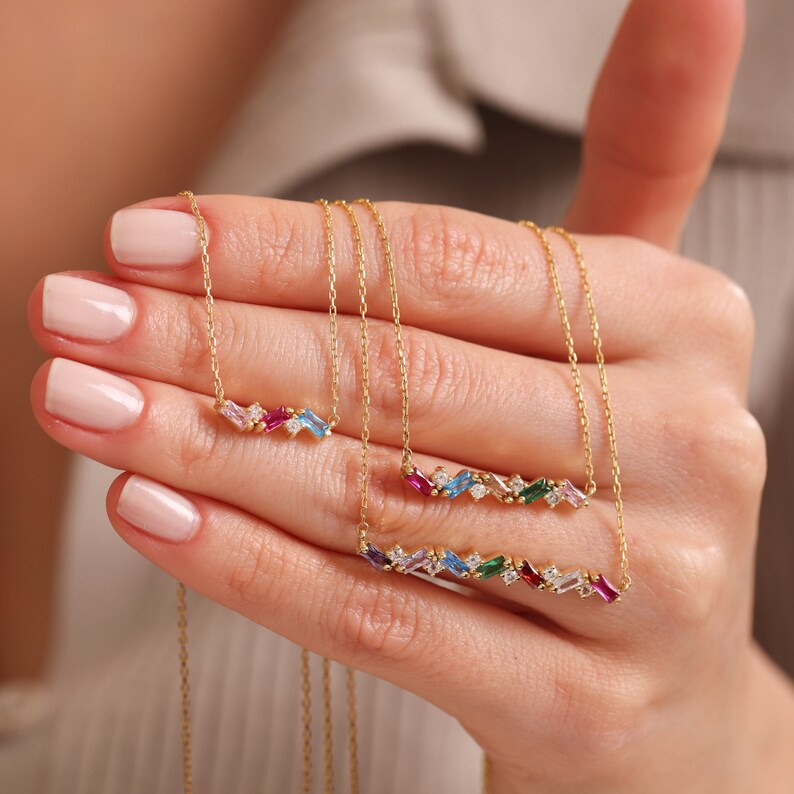 Puede incluir: Tres collares de oro con detalles de piedras preciosas de colores. Los collares presentan una cadena delicada y un colgante de barra con una fila de piedras preciosas en forma de baguette de varios colores, incluyendo rosa, azul, verde y rojo.