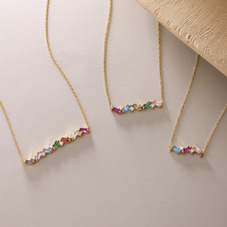 Puede incluir: Tres collares de oro con detalles de piedras preciosas de colores. Cada collar presenta una fila de peque&ntilde;as piedras preciosas rectangulares de varios colores, incluyendo rosa, azul, verde y rojo. Las piedras preciosas est&aacute;n engastadas en una cadena de oro.