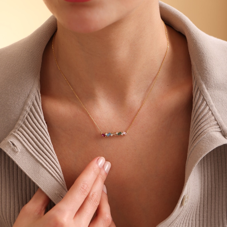 Puede incluir: Un collar de oro con un colgante de barra engastado con piedras preciosas de colores. Las piedras preciosas est&aacute;n dispuestas en fila e incluyen piedras rojas, rosas, azules, verdes y transparentes.