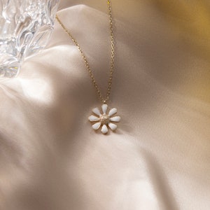 May include: A gold chain necklace with a white enamel daisy pendant. The necklace is on a light brown fabric background.