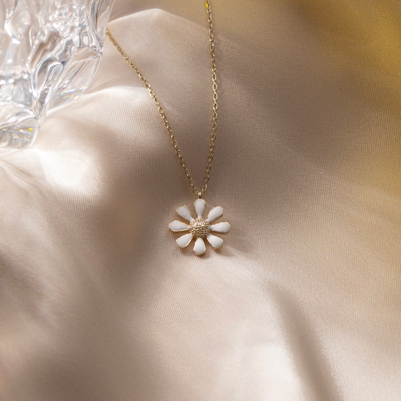May include: A gold chain necklace with a white enamel daisy pendant. The necklace is on a light brown fabric background.