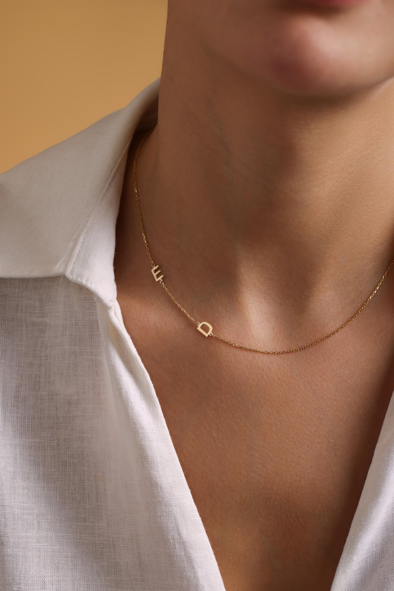 May include: A gold chain necklace with two small letters, "E" and "D", hanging from it. The necklace is worn by a person with a white shirt.