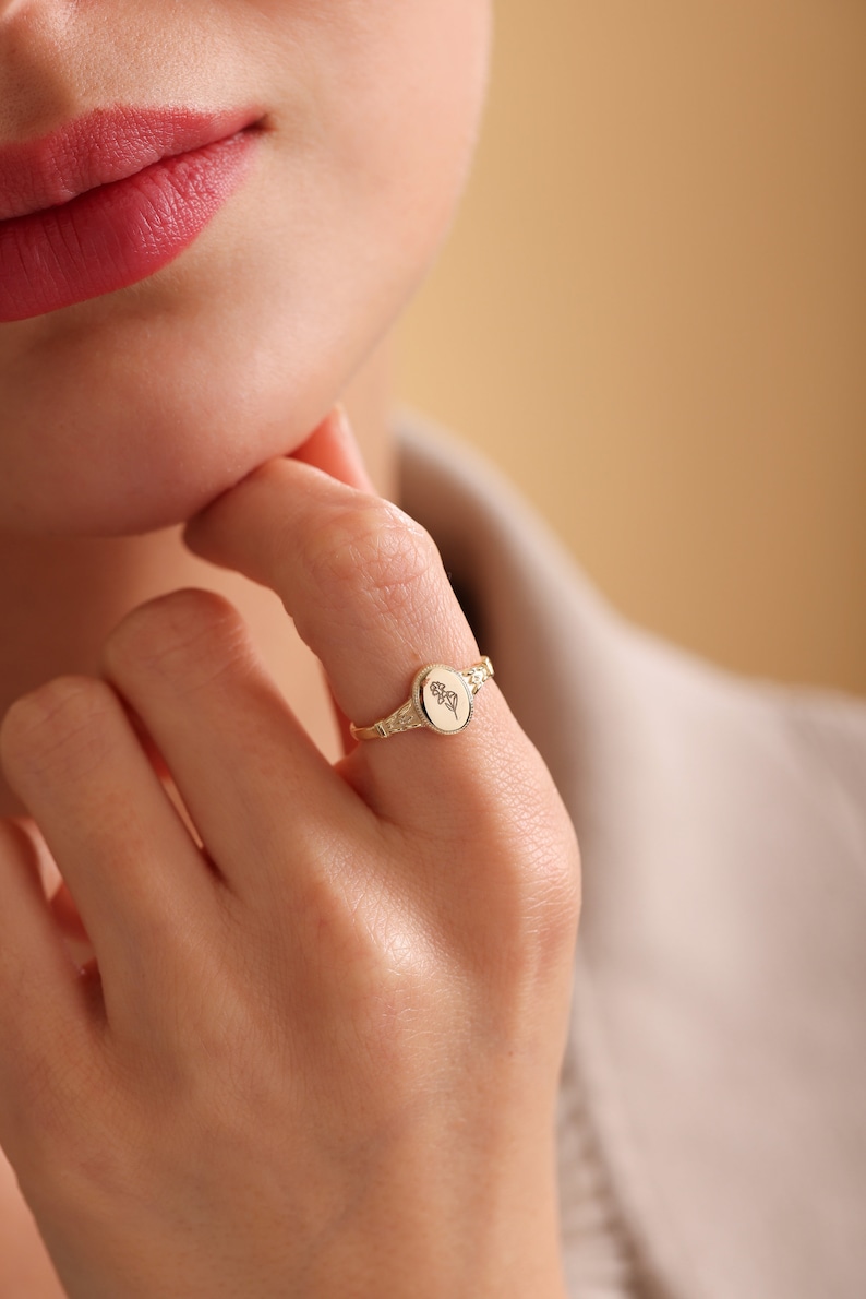May include: A gold ring with an oval-shaped setting featuring a delicate floral design. The ring is worn on the index finger of a person's hand.