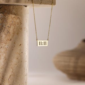 K&ouml;nnte beinhalten: Eine goldene Kettenkette mit einem rechteckigen Anh&auml;nger, auf dem "11:11" in Schwarz steht.