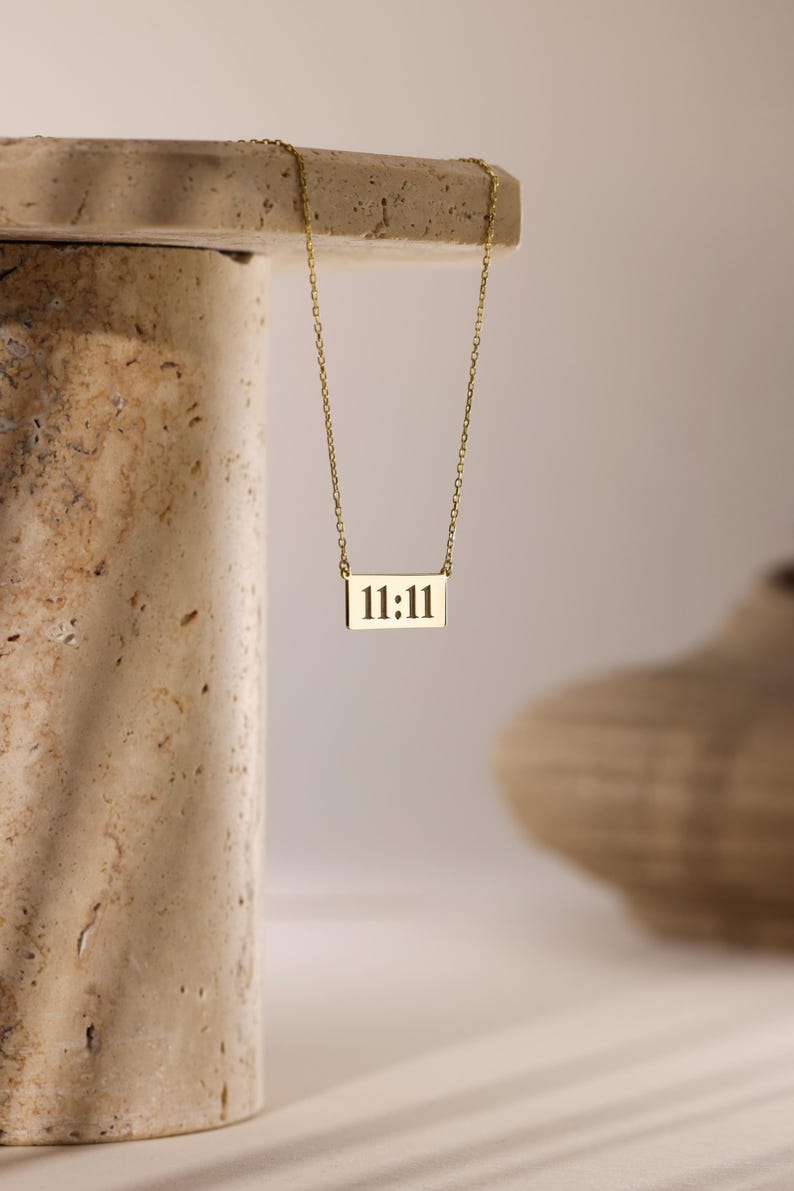 K&ouml;nnte beinhalten: Eine goldene Kettenkette mit einem rechteckigen Anh&auml;nger, auf dem "11:11" in Schwarz steht.