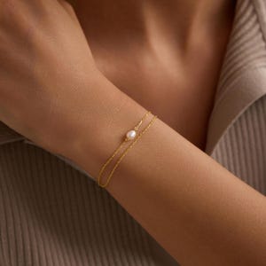 May include: A delicate gold bracelet featuring two fine chains, one adorned with a single, lustrous white pearl. The bracelet is worn on a wrist, showcasing its elegant simplicity. The jewelry is a classic accessory.