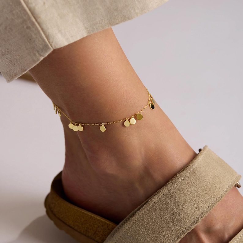 May include: A gold-colored anklet with small, round, dangling charms. The anklet is worn on an ankle with a sandal. The charms are evenly spaced along the delicate chain, creating a subtle, elegant look. The anklet is a piece of jewelry.