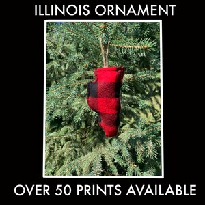 May include: A red and black plaid Illinois ornament hangs from a natural twine string against a backdrop of green pine needles. The ornament is shaped like the state of Illinois. Text at the top reads "ILLINOIS ORNAMENT" and at the bottom "OVER 50 PRINTS AVAILABLE."