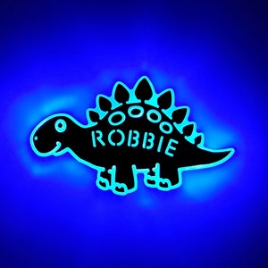 May include: A black dinosaur-shaped night light with the name "ROBBIE" on the back. The light is glowing blue.