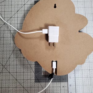 May include: The back of a wooden sign with a white electrical cord plugged into a white adapter. The cord is plugged into a USB port on the sign. The sign is on a grey grid surface.