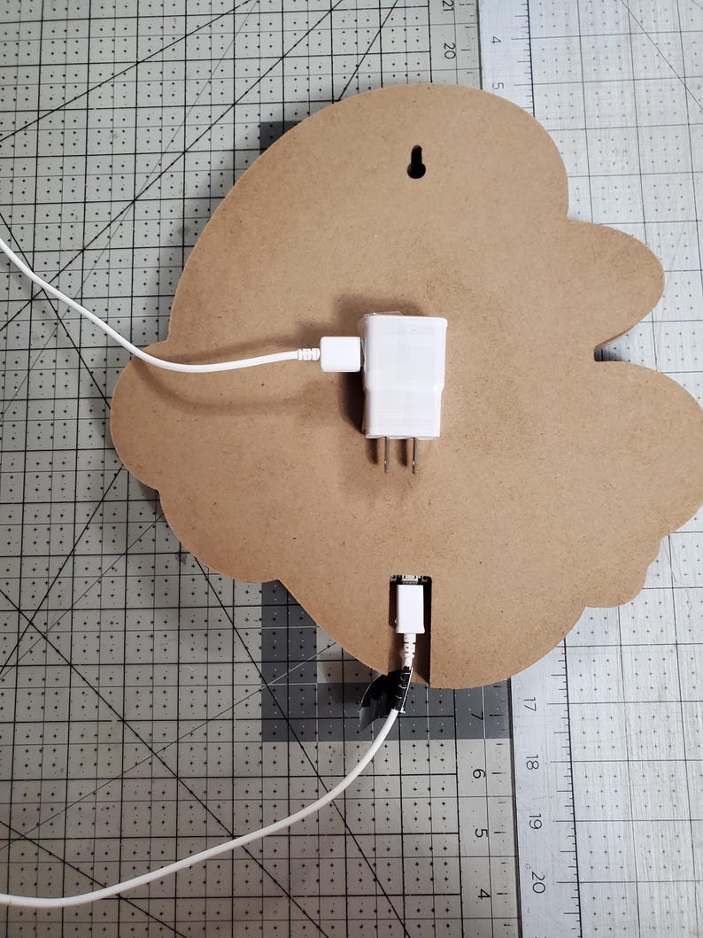 May include: The back of a wooden sign with a white electrical cord plugged into a white adapter. The cord is plugged into a USB port on the sign. The sign is on a grey grid surface.