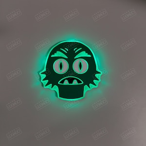 May include: A green, glowing, stylized creature face with large eyes and a wide mouth. The design is a cutout, with a bright green glow emanating from the edges. The product is likely a decorative item or a light-up sign.