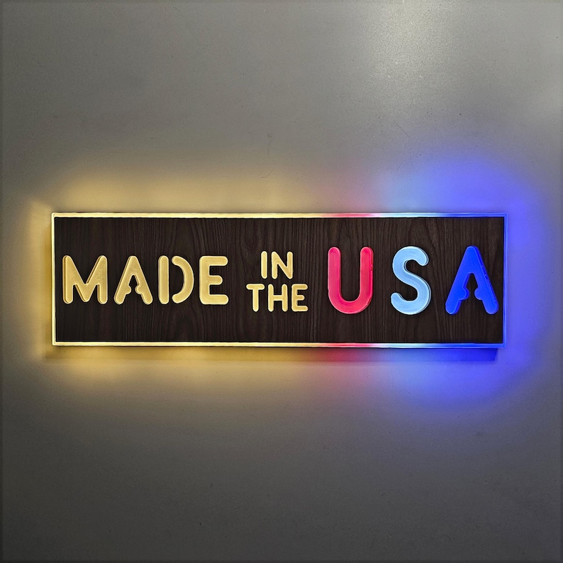 May include: A wooden sign with the text "MADE IN THE USA" in white letters. The letters "U" and "S" are illuminated in pink and blue, respectively.