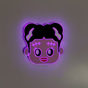 May include: A pink and black light-up wall decor featuring a cartoon girl with stitched eyebrows and a big smile. The light is purple.