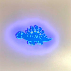 May include: A blue dinosaur night light with the name "ROBBIE" written on it. The light is glowing a bright purple colour.