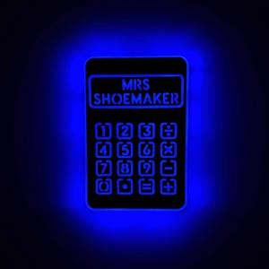 Puede incluir: Una calculadora negra con un brillo azul alrededor. La calculadora tiene el texto "MRS SHOEMAKER" en la pantalla superior y un teclado de calculadora estándar.