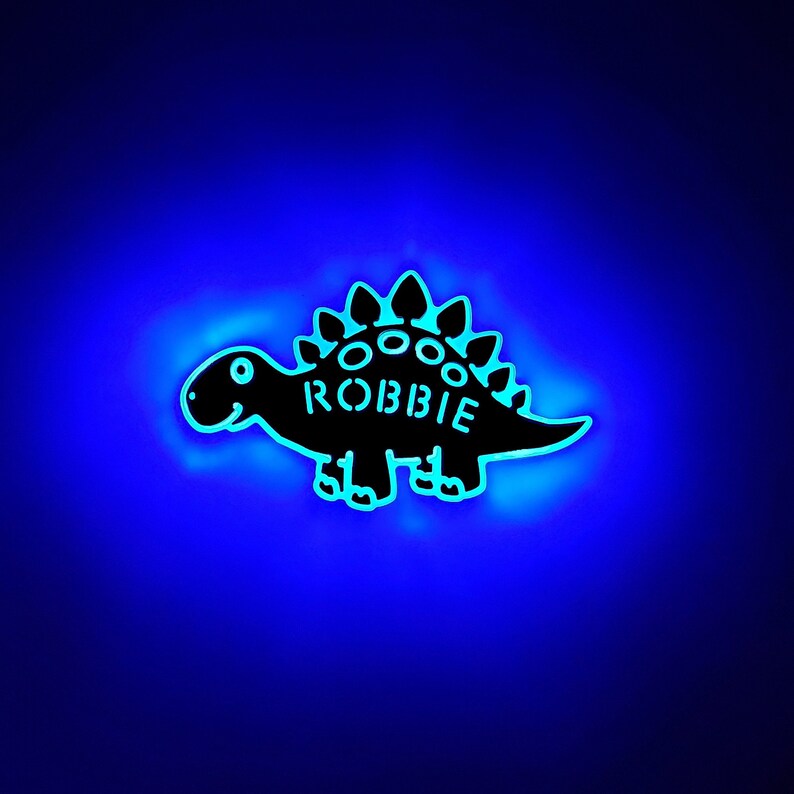 May include: A black dinosaur-shaped night light with the name "ROBBIE" on the back. The light is glowing blue.