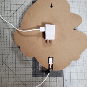 K&ouml;nnte beinhalten: Die R&uuml;ckseite eines Holzschildes mit einem wei&szlig;en Stromkabel, das in einen wei&szlig;en Adapter gesteckt ist. Das Kabel ist in einen USB-Anschluss am Schild gesteckt. Das Schild befindet sich auf einer grauen Gitteroberfl&auml;che.