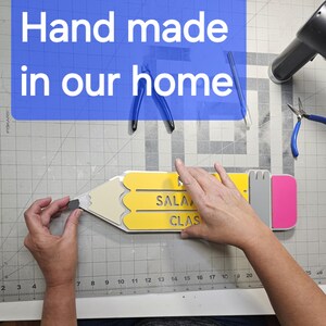 May include: A yellow and grey pencil-shaped wooden sign with the words "SALA" and "CLAS" cut out. The sign is being held by a person's hand. The sign is on a grey grid surface.