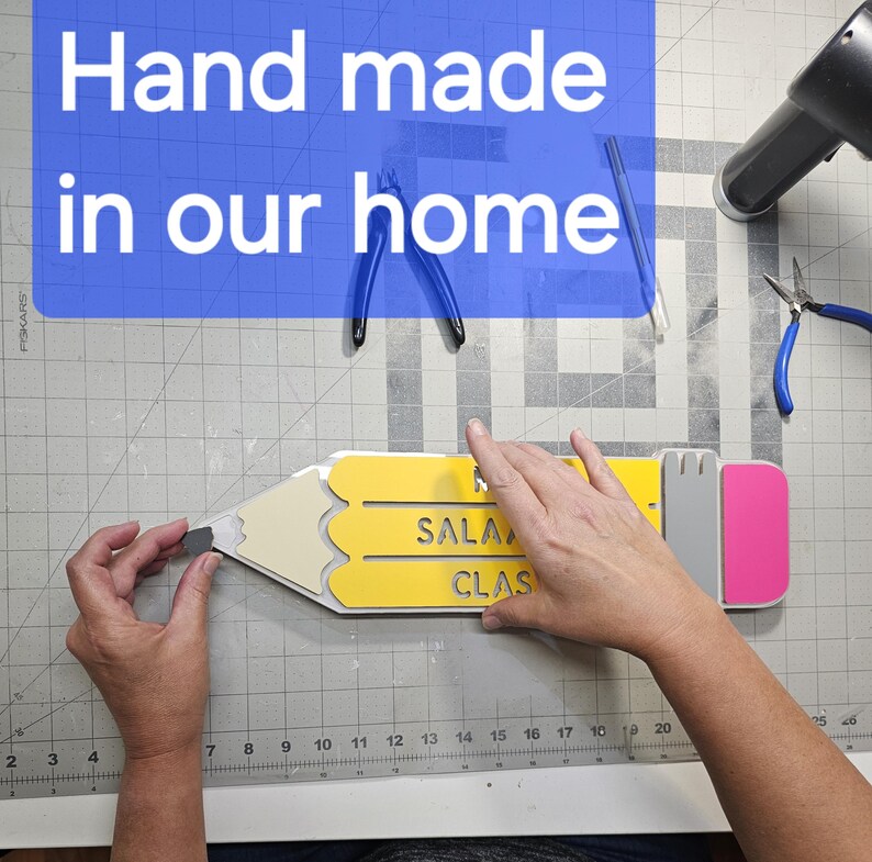 May include: A yellow and grey pencil-shaped wooden sign with the words "SALA" and "CLAS" cut out. The sign is being held by a person's hand. The sign is on a grey grid surface.
