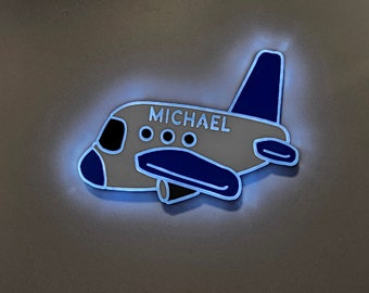 Puede incluir: Una luz nocturna de avión blanco con el nombre "MICHAEL" y cuatro pequeños círculos en el fuselaje. El avión está bordeado de azul y tiene una aleta de cola azul.