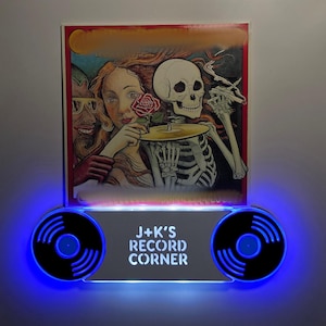 May include: A white acrylic record shelf with blue LED lights and black vinyl record silhouettes. The shelf has the text "J+K's Record Corner" printed on it. The shelf is mounted on a white wall and has a vintage record album with a skeleton and two women on it.