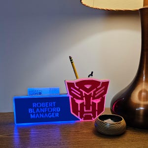 May include: A red and blue illuminated desk organizer with a Transformers logo. The organizer has a slot for business cards and a pencil holder. The text "ROBERT BLANFORD MANAGER" is displayed on the front of the organizer.