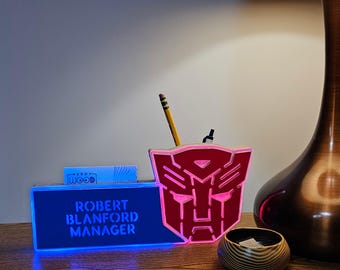 May include: A red and blue illuminated desk organizer with a Transformers logo. The organizer has a slot for business cards and a pencil holder. The text "ROBERT BLANFORD MANAGER" is displayed on the front of the organizer.