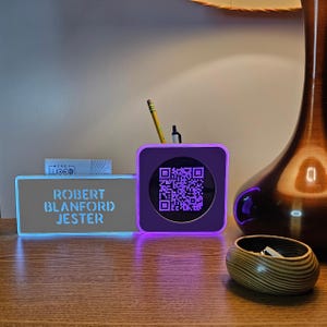 May include: A purple and blue illuminated desk organiser with a black and white QR code on the front. The organiser has a nameplate that reads "Robert Blanford Jester".