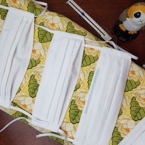 Pode incluir: Quatro máscaras de tecido branco com pregas são colocadas em um tecido floral amarelo e verde. As máscaras têm elásticos brancos.