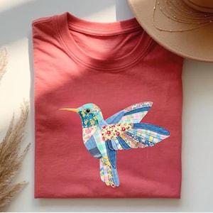 Op de afbeelding: Een koraalkleurig T-shirt met een gedetailleerde applicatie van een kolibrie. De kolibrie heeft een patchwork-ontwerp met blauwe, bloemen- en denimstoffen. De vogel heeft een gele snavel en is gecentreerd op het opgevouwen shirt.